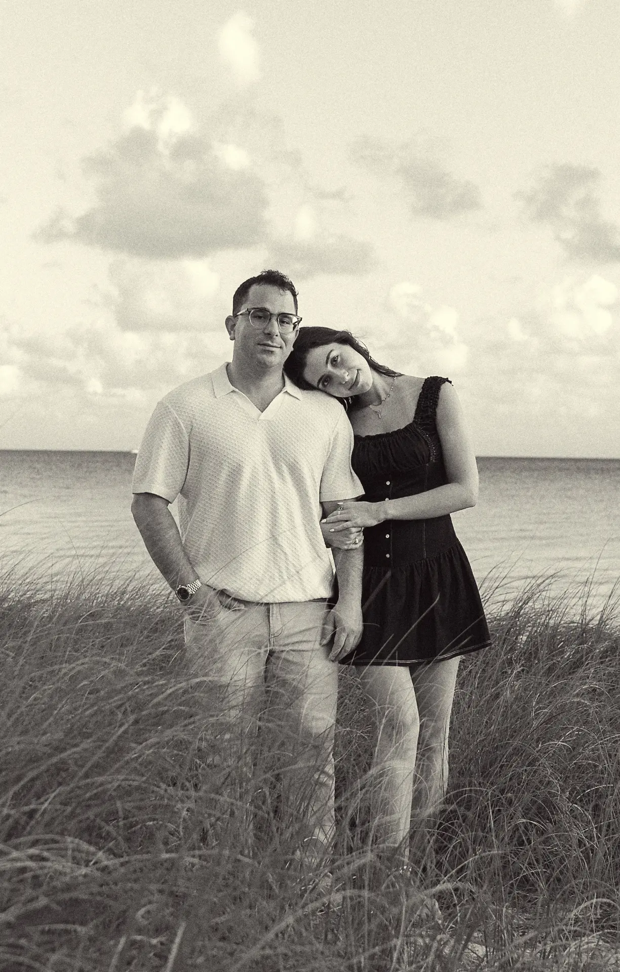 a couple in tall grass by the water, her head resting on his shoulder